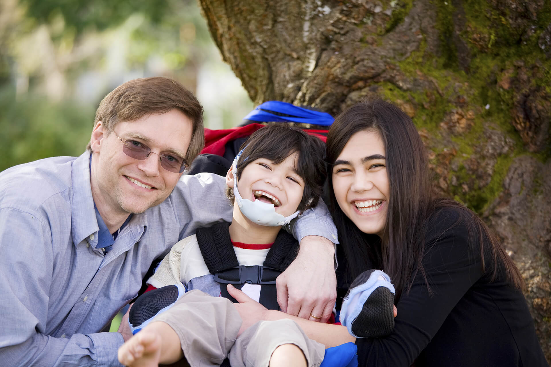 Happy disabled boy with cerebral palsy in wheelchair surrounded by father and older sister, laughing
